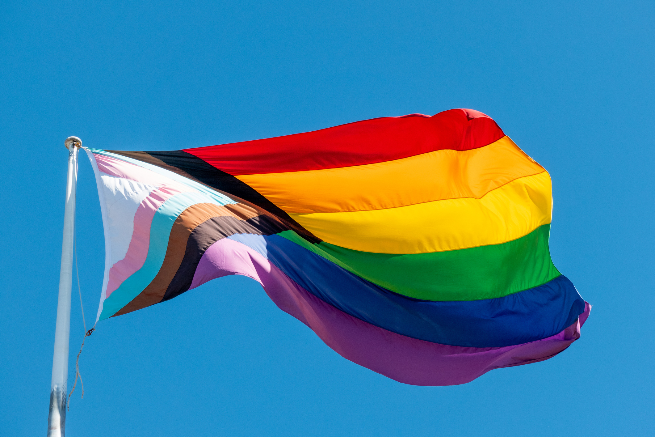 Rainbow Pride flag streaming in the wind against a clear blue sky on a flagpole; vibrant colors ripple in the breeze.