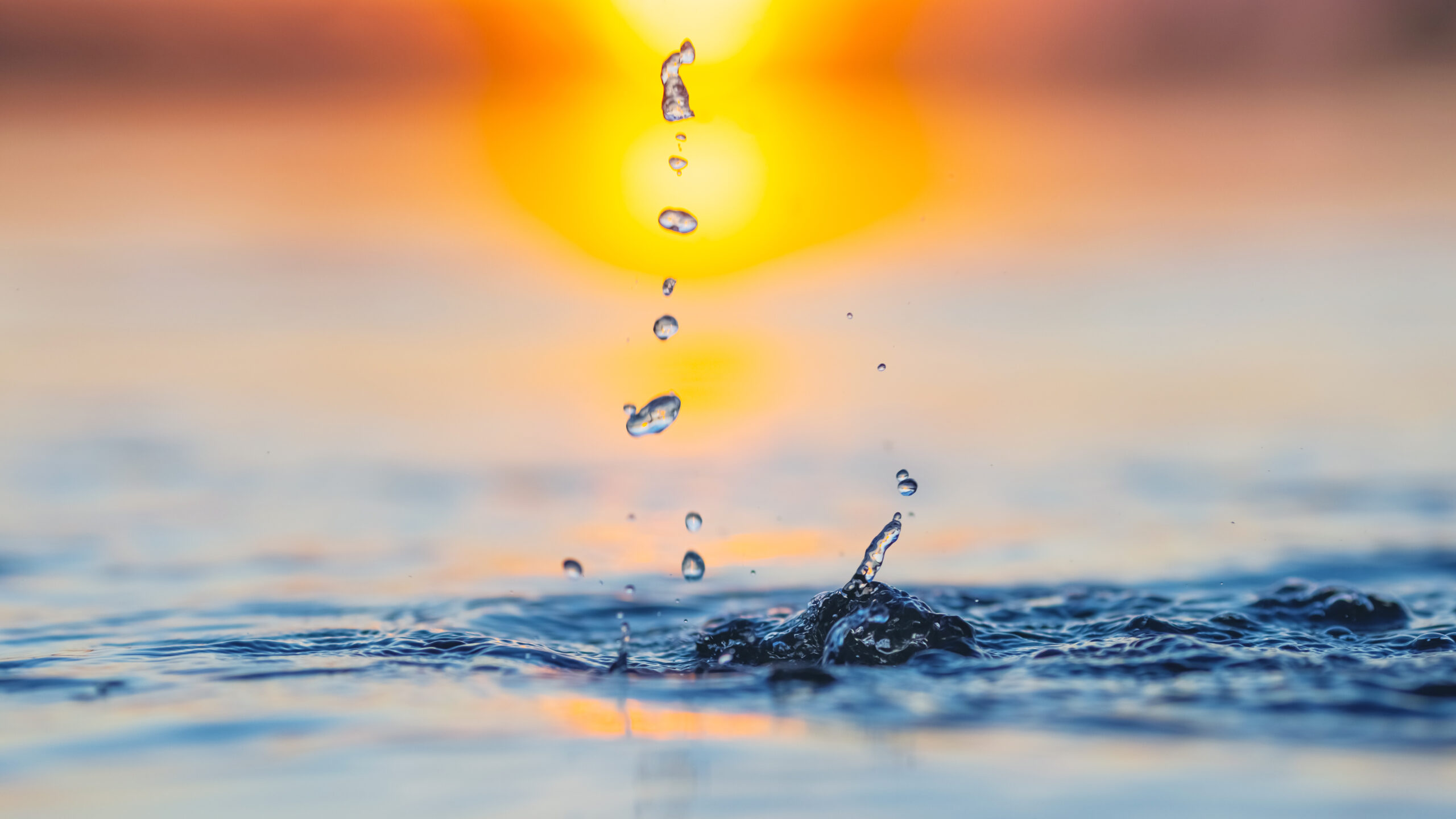 Water droplets midair above a calm sea at sunset with a warm orange and yellow sky.