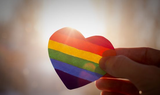 Close-up of a rainbow-striped heart held by fingers against a bright, blurred background, symbolizing LGBTQ+ pride.