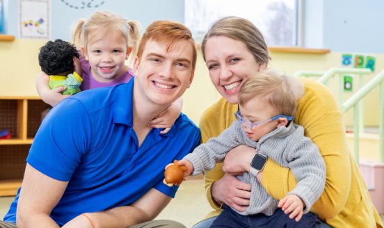 Smiling family in a bright playroom: father, mother, and two young children.