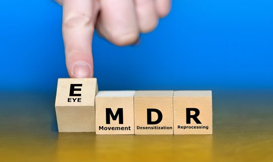 Close-up of wooden blocks spelling EMDR with Movement, Desensitization, Reprocessing; a finger taps the E block on the left.