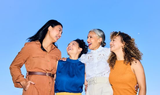 Four women of diverse ages smiling and chatting, arms around each other against a clear blue sky.