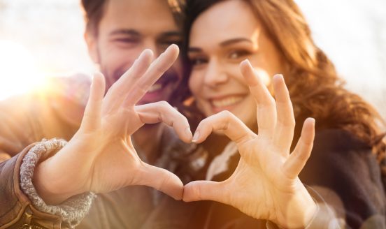 Couple smiling and forming a heart shape with their hands in warm sunlight in the foreground.