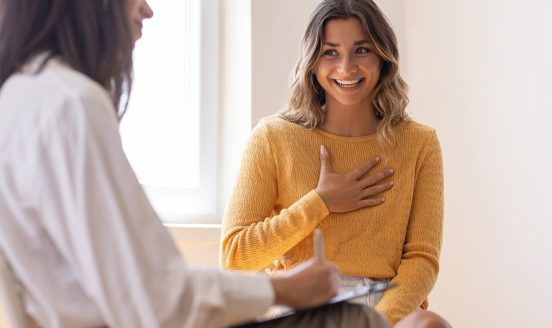 A psychologist listening as a smiling client places a hand on her chest during a therapy session